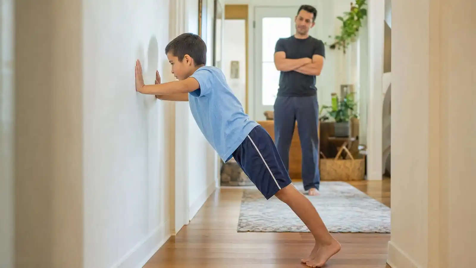A child doing wall push-ups in a bright hallway for calming proprioceptive input while a parent watches supportively from a few steps back